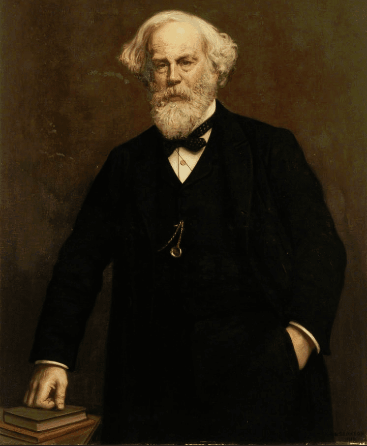 Man with white hair and beard, dressed in suit with watch chain and bowtie, leaning on books.