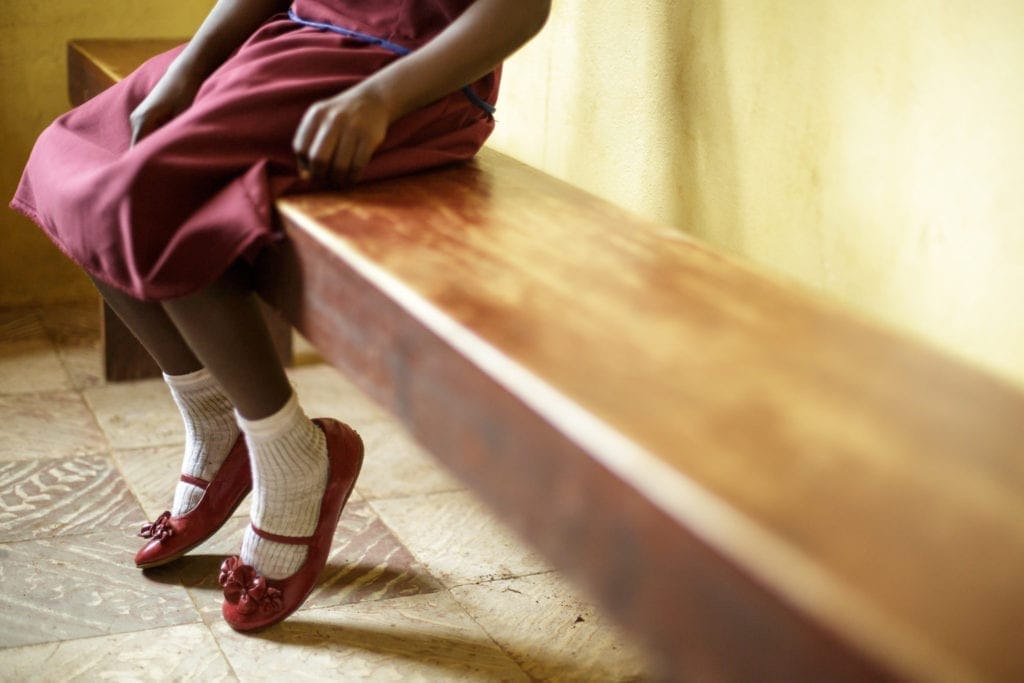 Lower body and legs of dark-skinned girl sitting on bench, as though in waiting at a clinic. "6 February 2014. International Day of Zero Tolerance of Female Genital Mutilation." UNICEF / Olivier Asselin via MONUSCO Photos on Flickr. CC BY-SA 2.0.