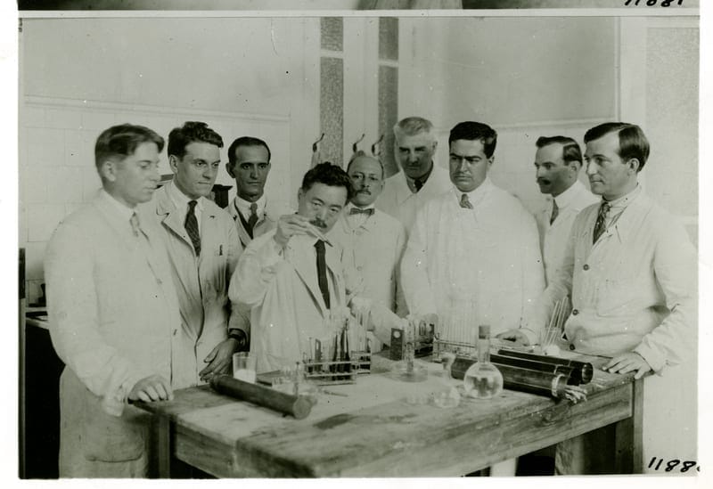 Black and white photo of nine men in lab coats around a laboratory table with Hideyo Noguchi, who researched the cause of yellow fever, left of center, holding up a test tube as if demonstrating to the others.