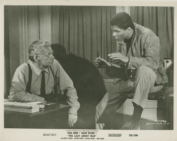 Man with glasses and mustache in suspenders, seated, listening to another man, seated across from him