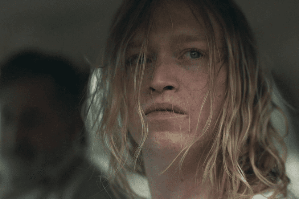 Young man with disheveled blond hair looking out car window as a man with a mustache in the blurred background behind him looks at him