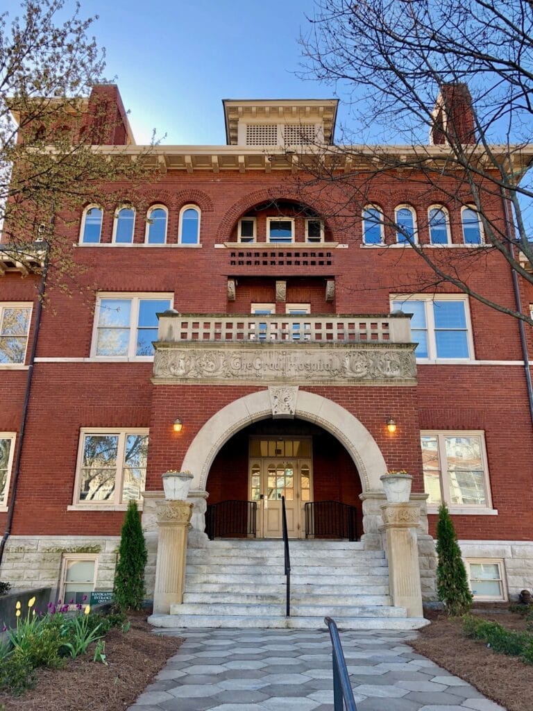 Photo of the Old Grady Memorial Hospital main building, now known as Georgia Hall, a three-story red-brick building with a double-wide staircase and white stone accents (such as arches) in its design. "The Grady Hospital" is carved into the white stone above the entrance.